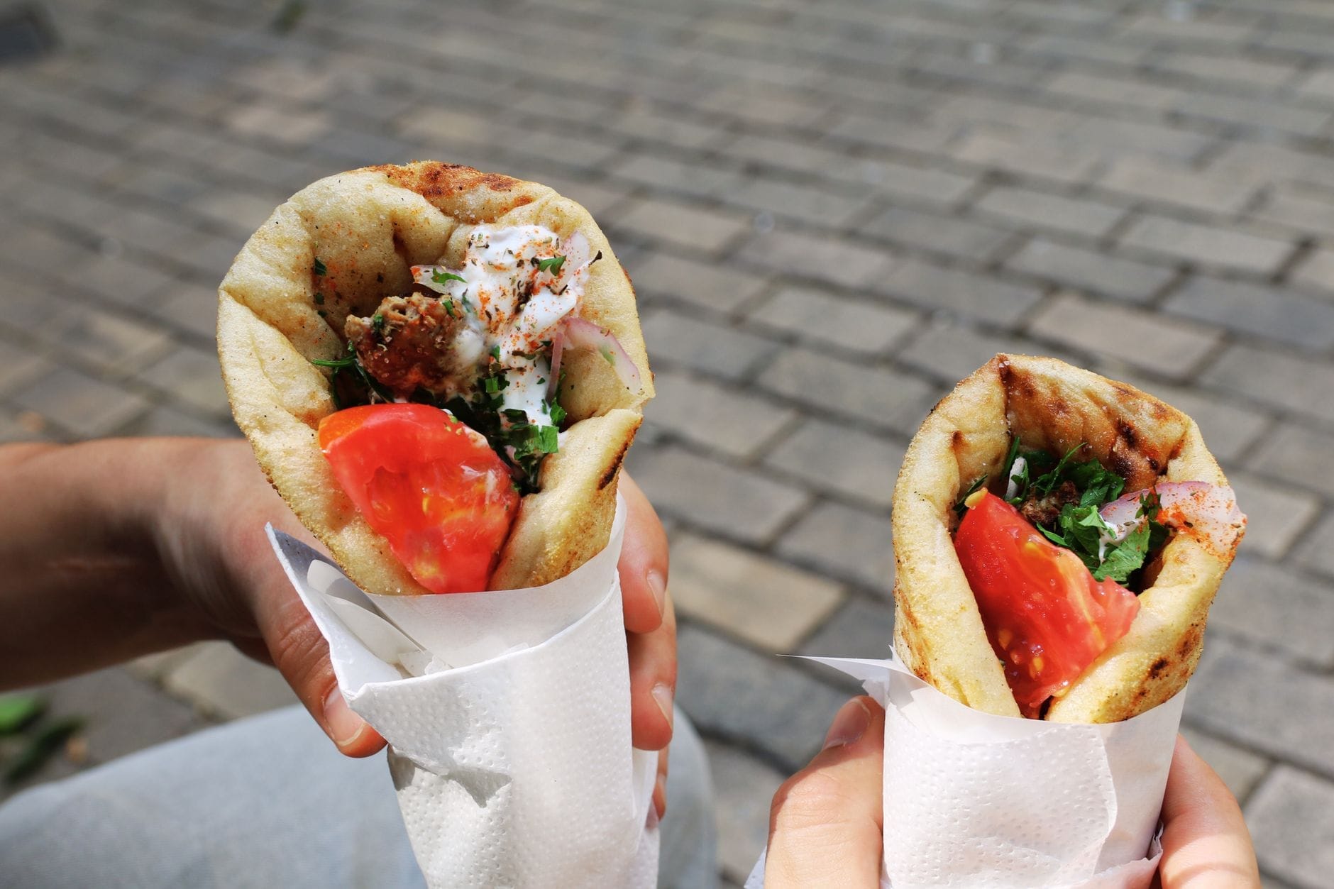 Due mani che tengono pita gyros avvolti nella carta, ripieni di carne, pomodori, erbe aromatiche e salsa, con uno sfondo di pavimentazione in pietra.