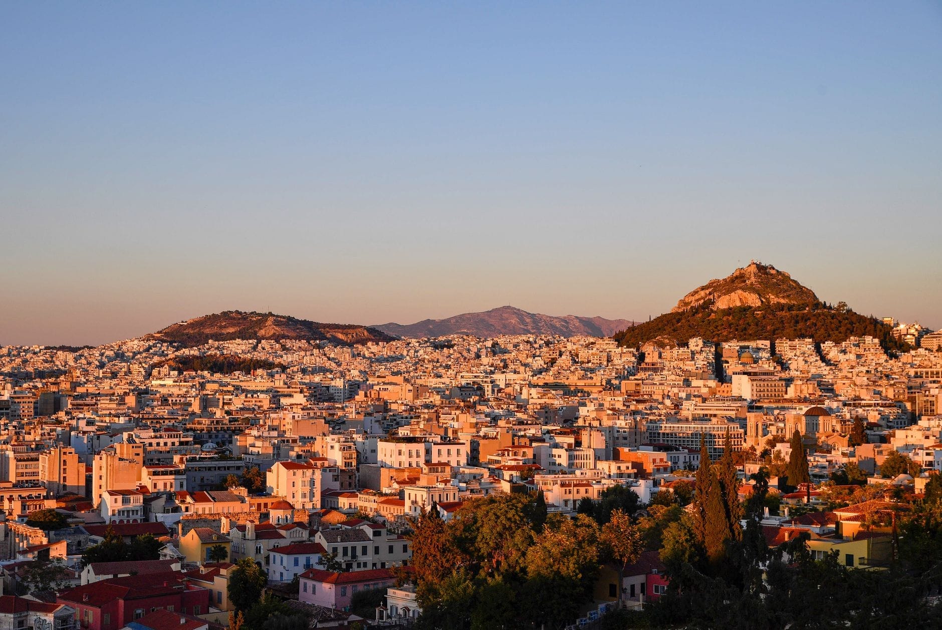 Un ampio e colorato paesaggio urbano di Atene al tramonto con il Monte Licabetto sullo sfondo e densi edifici urbani in primo piano.