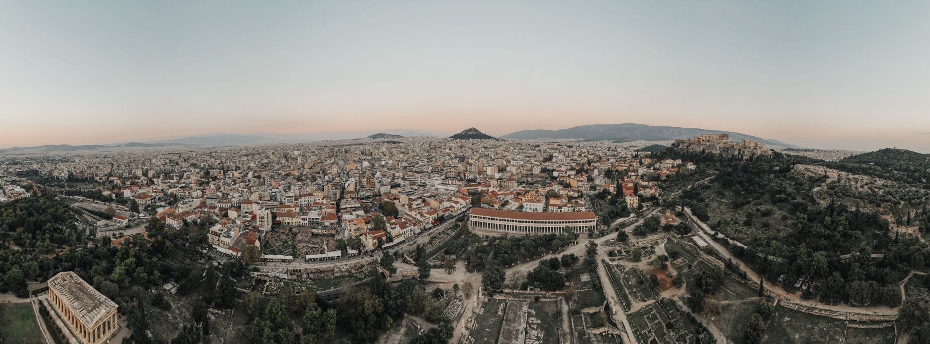 Vista aérea panorâmica de Atenas ao amanhecer com a Acrópole e os bairros ao redor, mostrando a escala e a paisagem da cidade sob um céu suave e calmo.