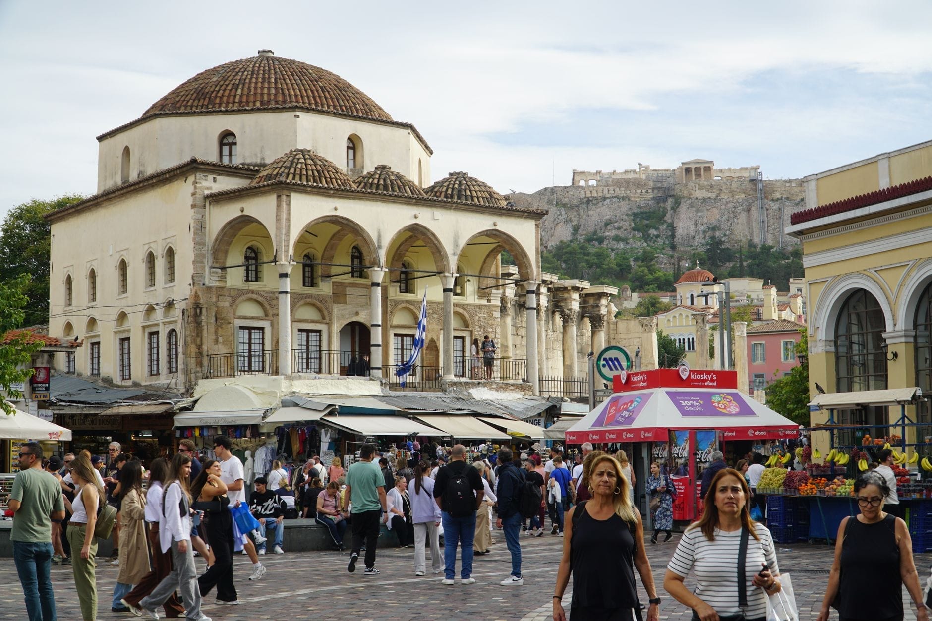 Concurrida plaza de Monastiraki en Atenas con personas, puestos de mercado, una mezquita histórica y la Acrópolis al fondo en un día soleado.