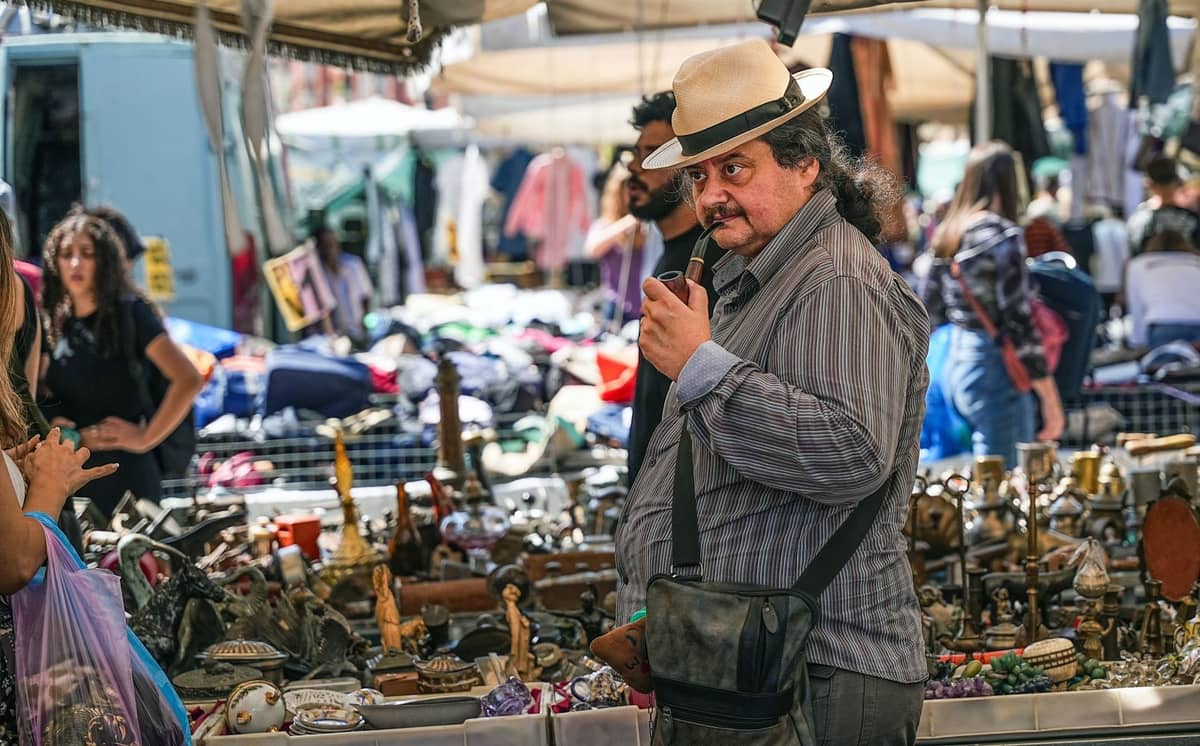 Hombre con sombrero examinando pequeñas antigüedades en un puesto de mercadillo al aire libre repleto de diversos artículos de segunda mano y personas curioseando.