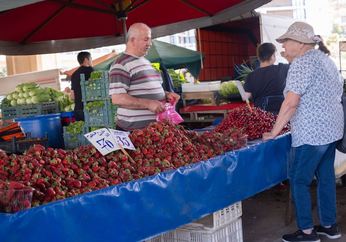 Escena de mercado de agricultores al aire libre en Atenas con un vendedor que ofrece fresas frescas y otros productos a una mujer bajo una carpa.