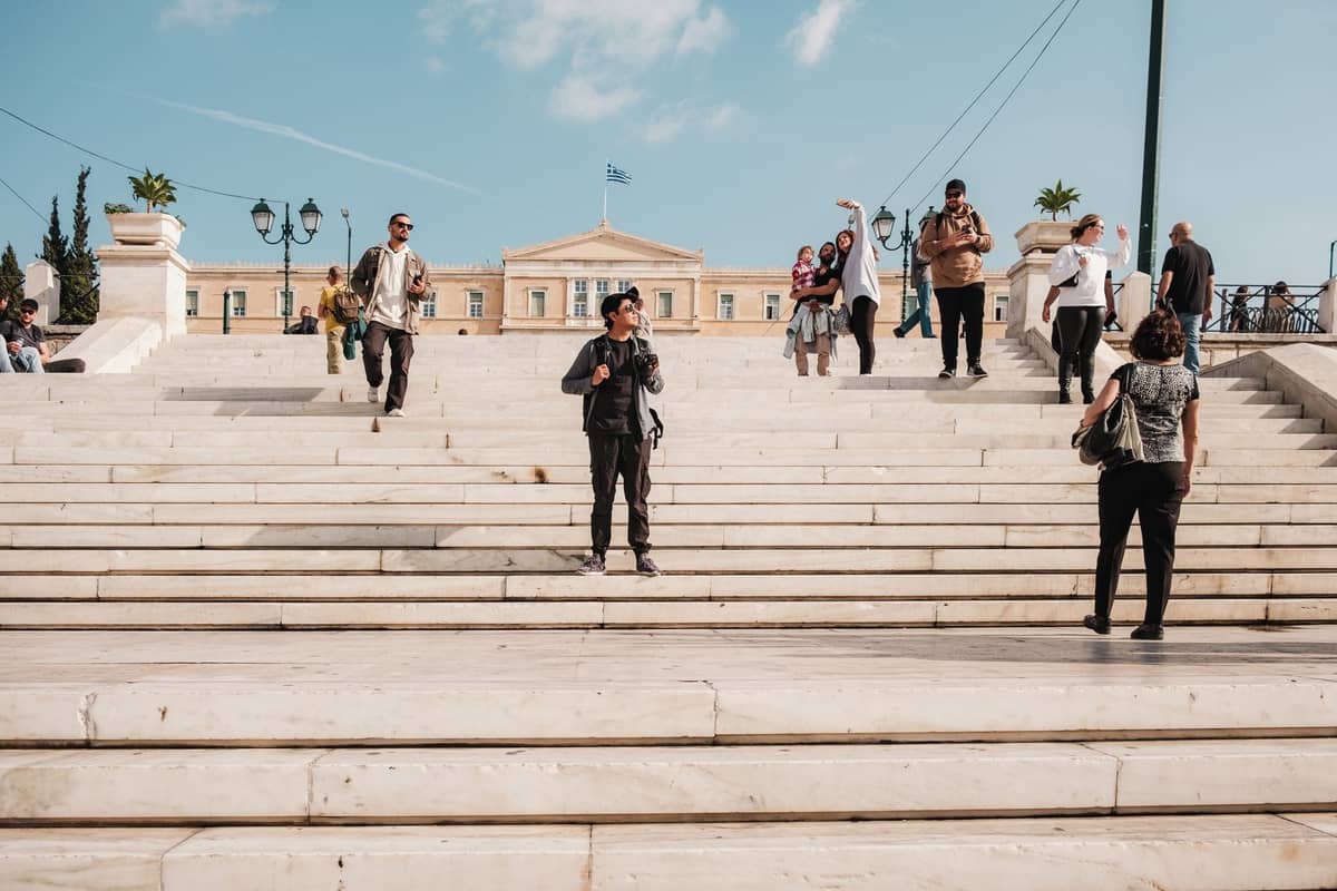 Touristen spazieren und fotografieren auf den Marmorstufen des Syntagma Square mit dem Gebäude des Hellenischen Parlaments im Hintergrund unter einem blauen Himmel.