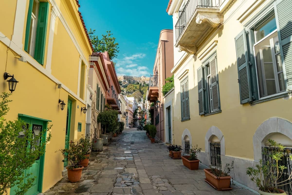 Charmante Fußgängerzone in Athen mit bunten Häusern, Topfpflanzen und der Akropolis im Hintergrund unter einem blauen Himmel.