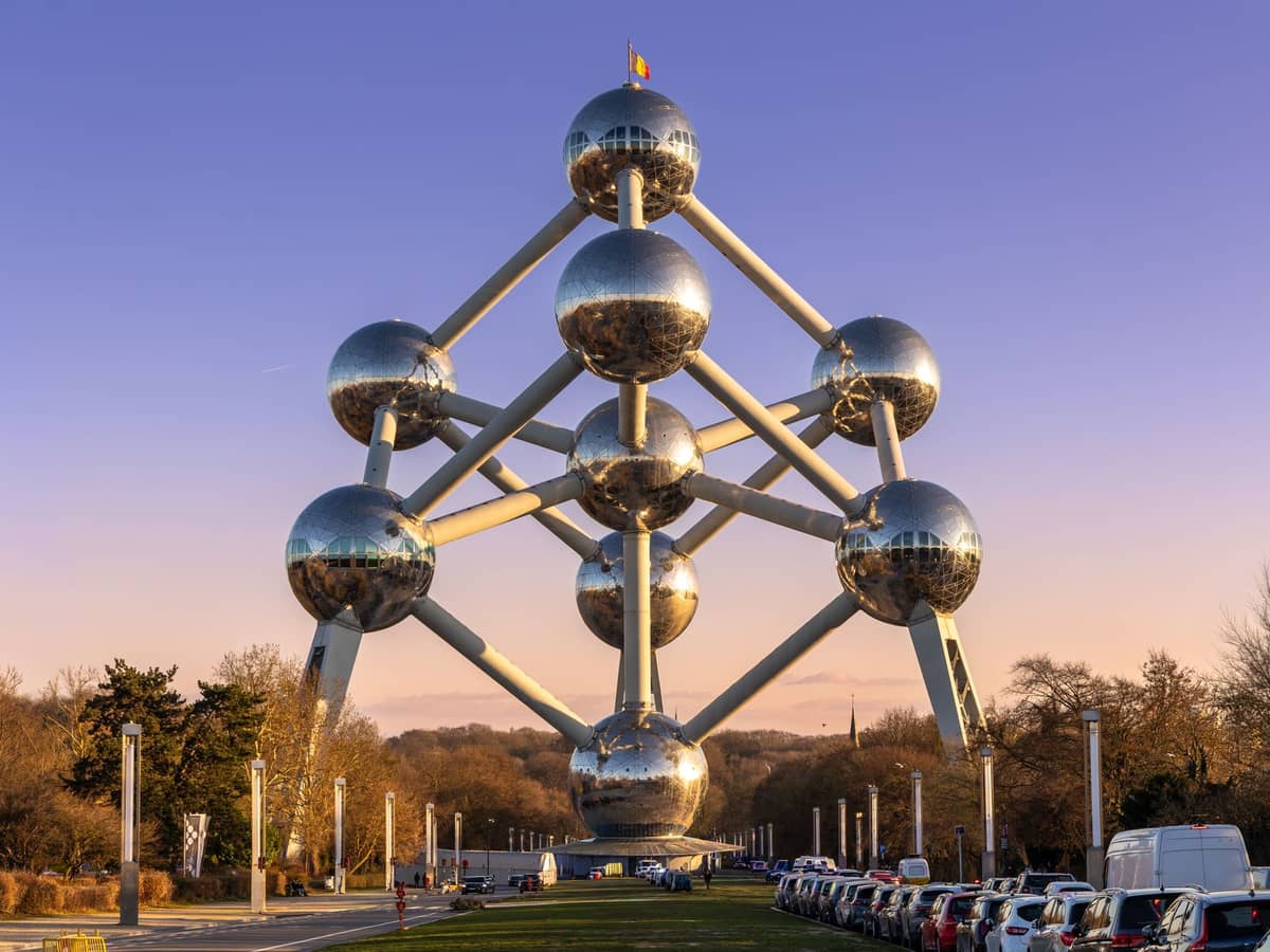 Vista completa da estrutura do Atomium em Bruxelas ao pôr do sol, mostrando todas as nove esferas de aço inoxidável conectadas por tubos, rodeadas de árvores e carros estacionados.