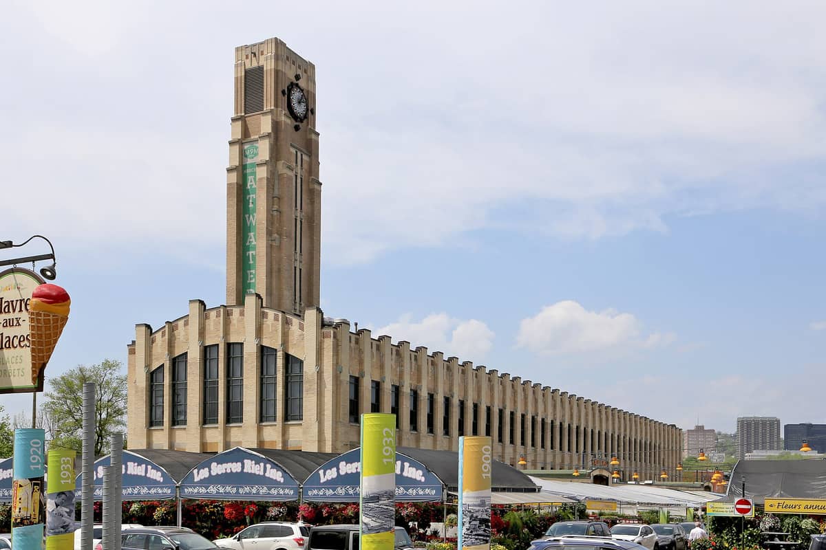 Außenansicht des Atwater Market in Montreal mit seinem markanten Art-Déco-Uhrturm, Marktständen und bunten Blumen unter blauem Himmel.