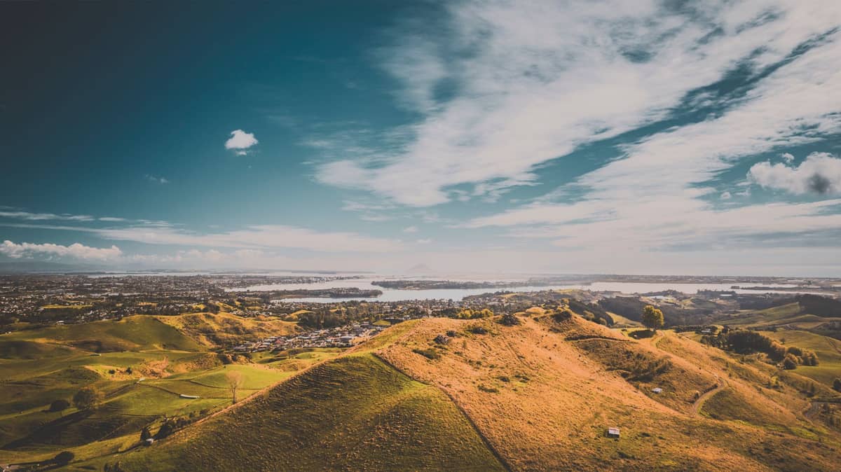 Weiter Blick auf vulkanische Hügel und grasbewachsene Kämme mit Blick auf Auckland, im Hintergrund die Stadt und der Hafen unter einem dramatischen Himmel.