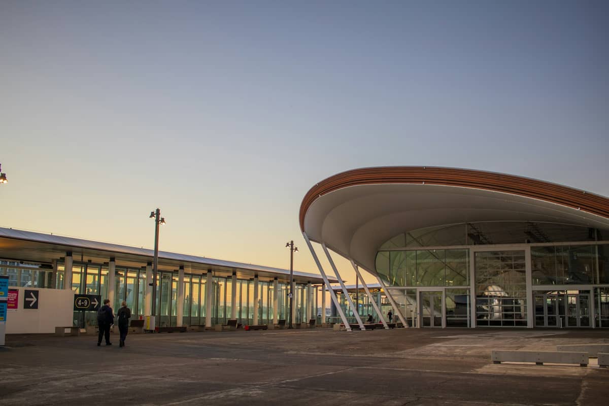 Außenansicht des Auckland Airport in der Abenddämmerung mit dem modernen geschwungenen Terminaleingang aus Glas und zwei Personen davor.