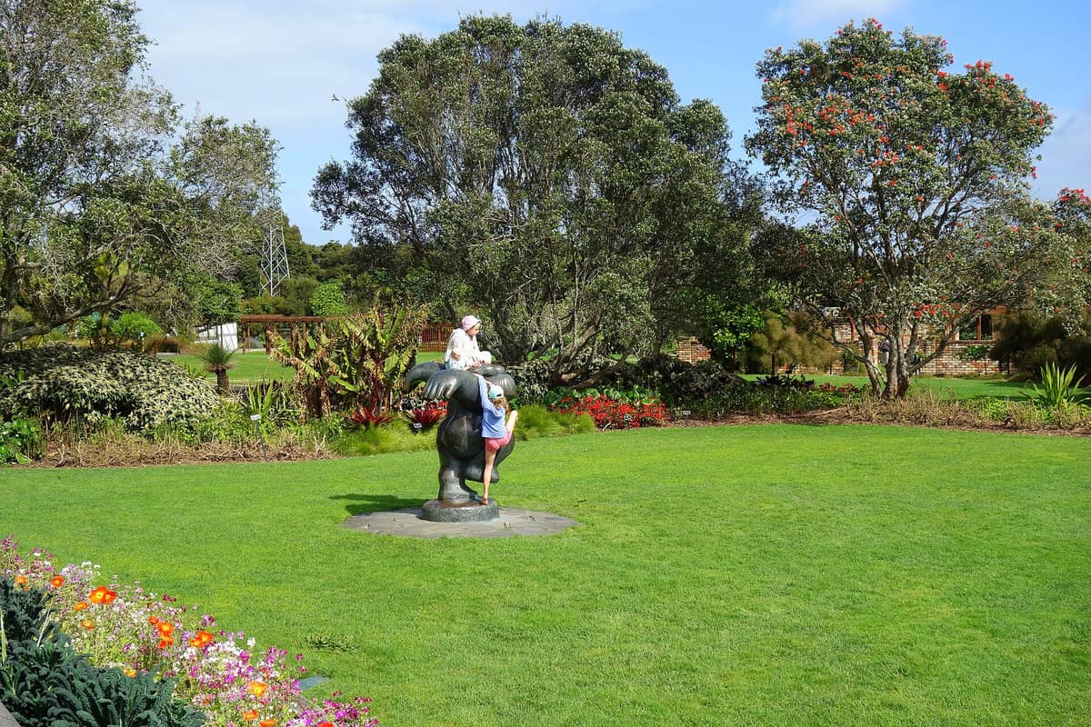 Weitläufiger Blick über die Auckland Botanic Gardens mit grüner Rasenfläche, bunten Blumenbeeten, üppigen Bäumen und einer verspielten Skulptur, auf der Kinder klettern.