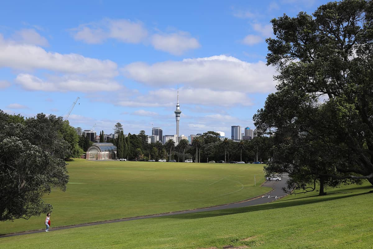 Weiter Blick über die offenen Rasenflächen des Auckland Domain mit dem Wintergarden-Glashaus, Palmen und der Skyline Aucklands einschließlich des Sky Tower in der Ferne.