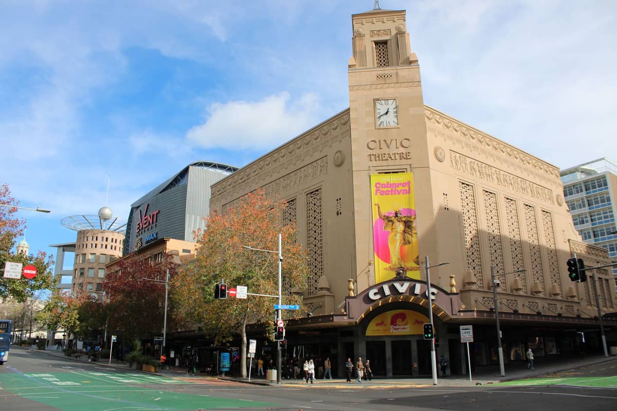Widok ulicy w ciągu dnia na Civic Theatre w Auckland, kultowe miejsce sztuk scenicznych w stylu art déco, z kilkoma przechodzącymi osobami i banerem reklamującym festiwal.