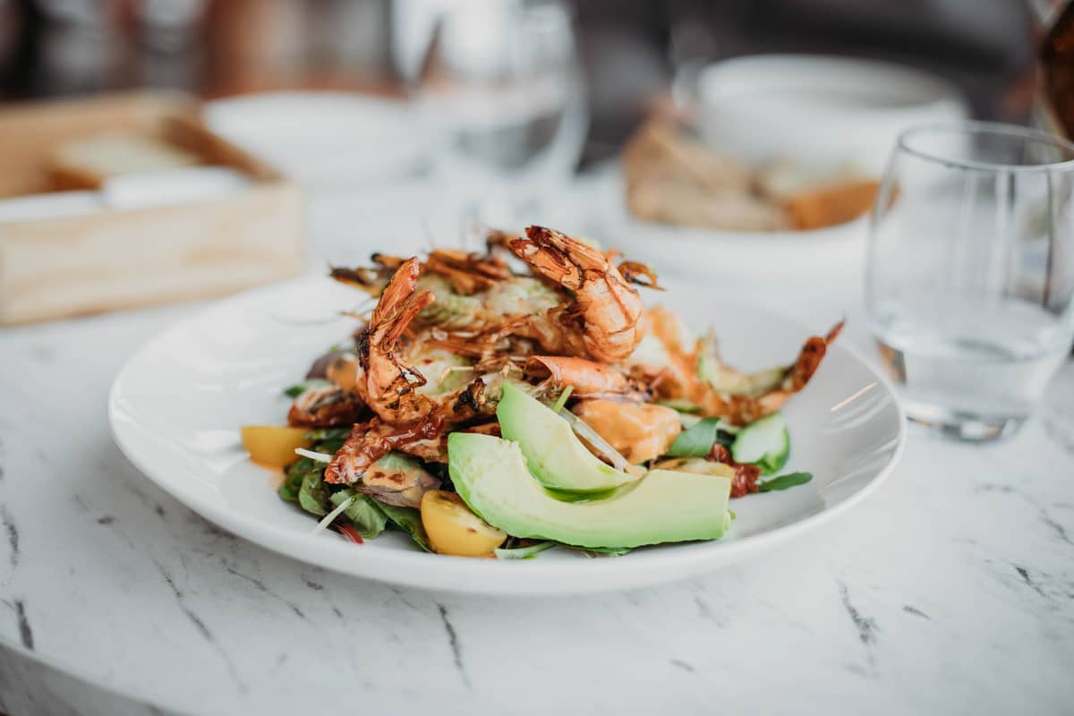 Ein Teller mit frischem Meeresfrüchtesalat mit gegrillten Garnelen, Avocado und Gemüse auf einem weißen Teller an einem Restauranttisch.