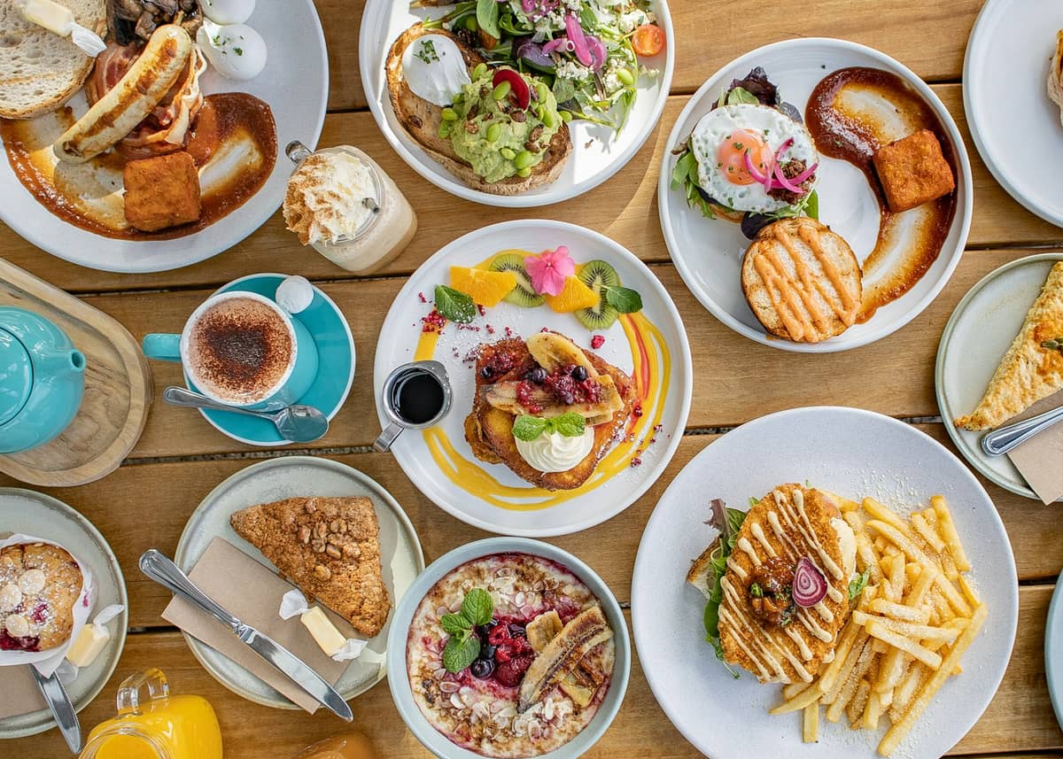Eine Auswahl an Brunch-Gerichten und Getränken auf einem Holztisch, darunter Eggs Benedict, Toast, Salat, Flat Whites, Saft und Café-Servings.