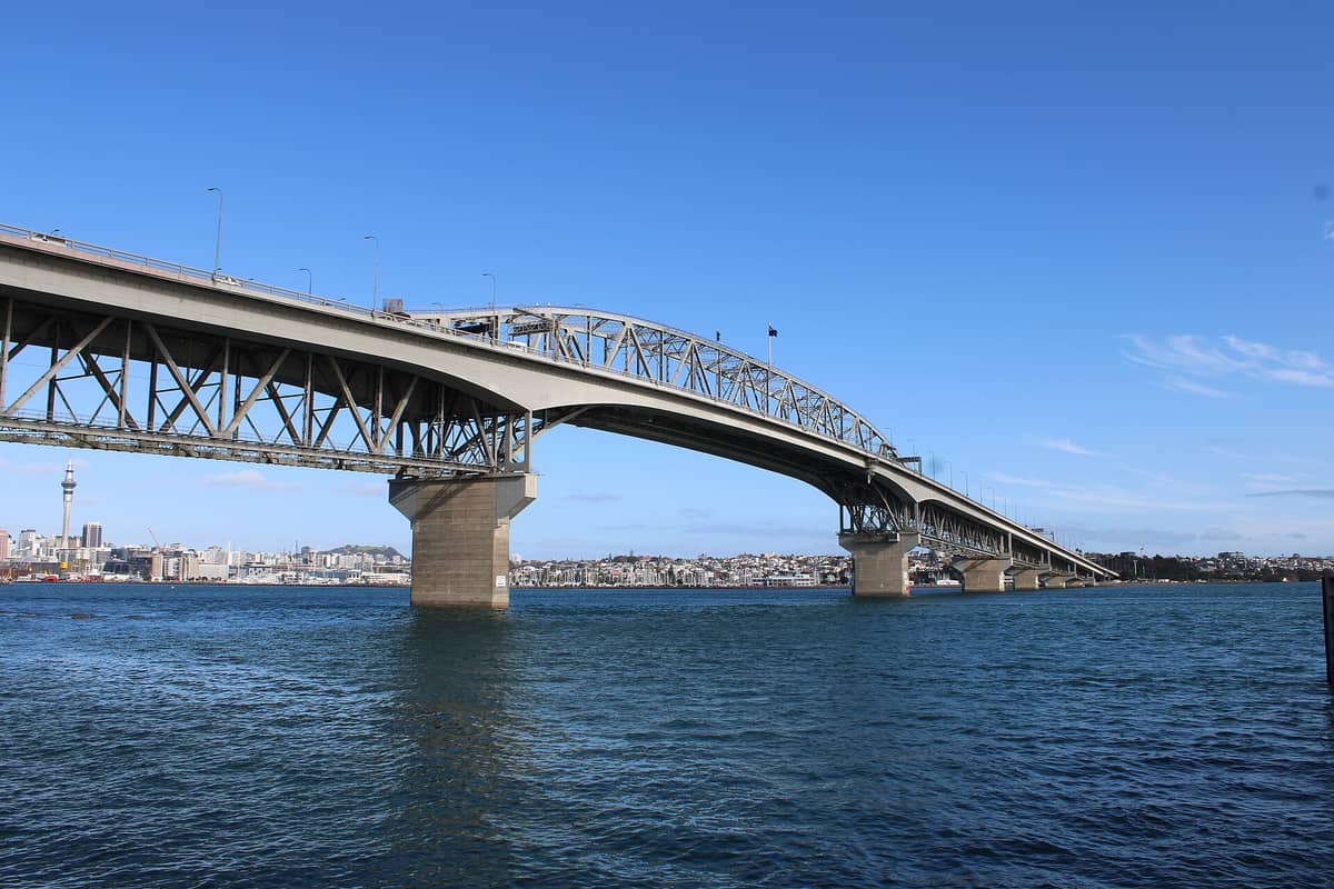 Breite Tagesansicht der Auckland Harbour Bridge vom Wasser aus, mit der Skyline der Stadt, blauem Himmel und dem Waitematā Harbour im Hintergrund.