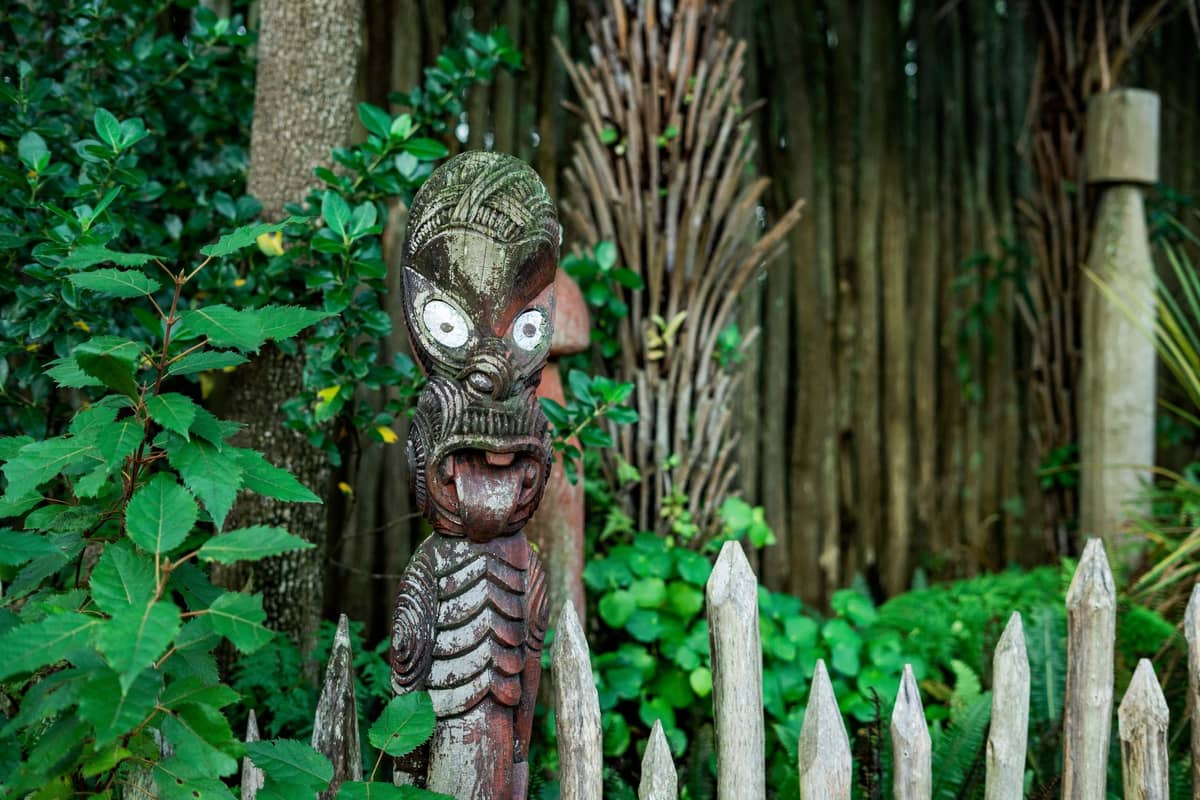 A traditional Māori carved wooden statue stands beside a wooden fence and native plants in a lush outdoor setting.