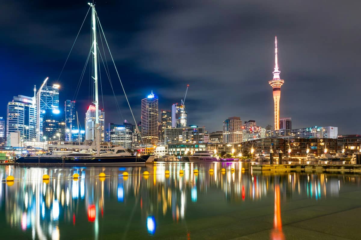 Frente marítimo del CBD de Auckland de noche, con rascacielos iluminados, Sky Tower y reflejos coloridos en el puerto con un yate atracado.
