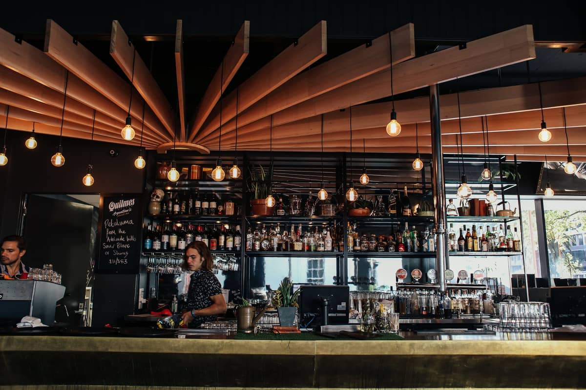 Interior de bar contemporáneo con iluminación elegante, botellas en estantes y bartenders preparando bebidas detrás del mostrador en un ambiente animado.