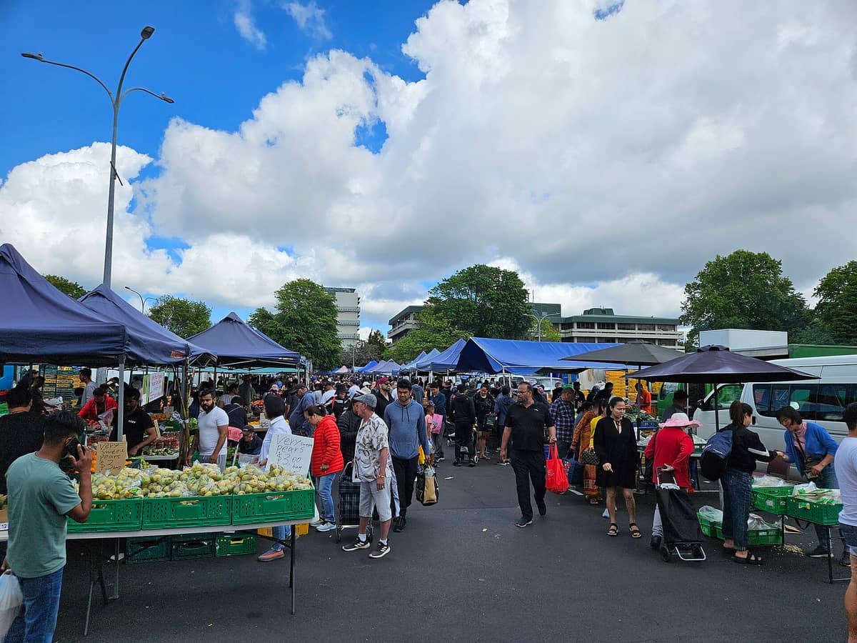 Scène de marché en plein air à Auckland avec des personnes diverses faisant leurs courses aux étals de nourriture et de produits sous des tentes bleues par une journée ensoleillée.