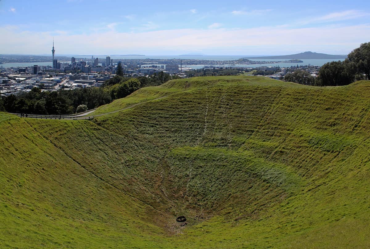 Vue panoramique du cratère volcanique herbeux de Mount Eden avec la skyline d'Auckland et le port en arrière-plan par temps clair.