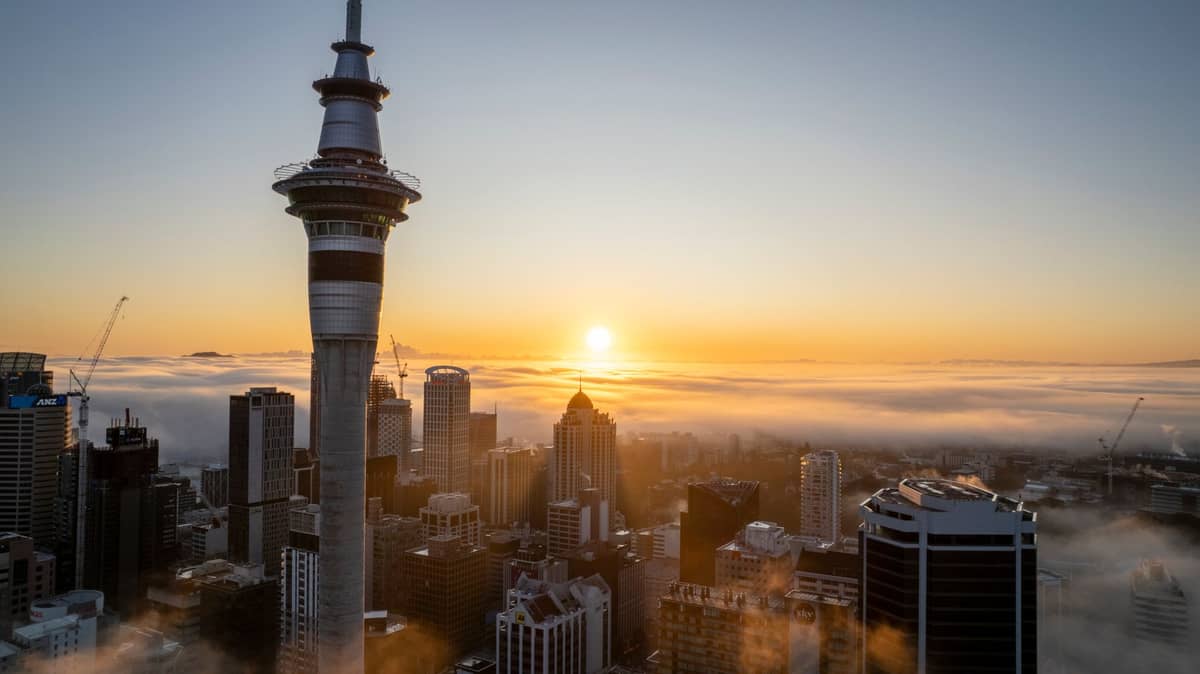 Ein breites Panoramafoto der Skyline Aucklands bei Sonnenaufgang – der Sky Tower im Vordergrund, goldenes Licht strahlt durch tief hängende Wolken.