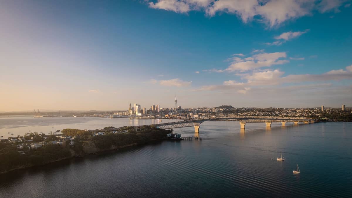 Ein weitläufiges Luftbild der Skyline von Auckland mit dem Sky Tower und der Auckland Harbour Bridge, im Vordergrund Boote und Wasser unter einem teils bewölkten Himmel.