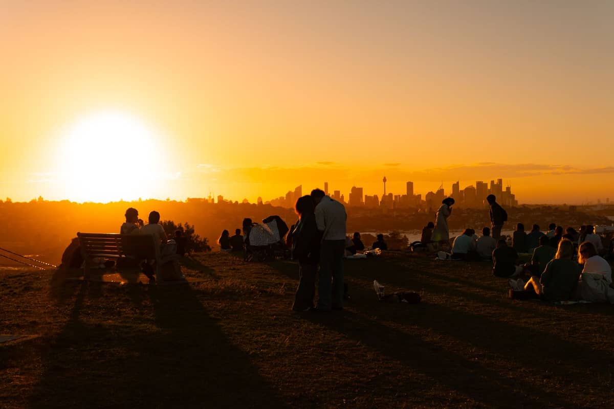 Grande gruppo di persone riunite su una collina erbosa al tramonto con vista sullo skyline di Auckland, a illustrare le folle stagionali sulle vette vulcaniche.