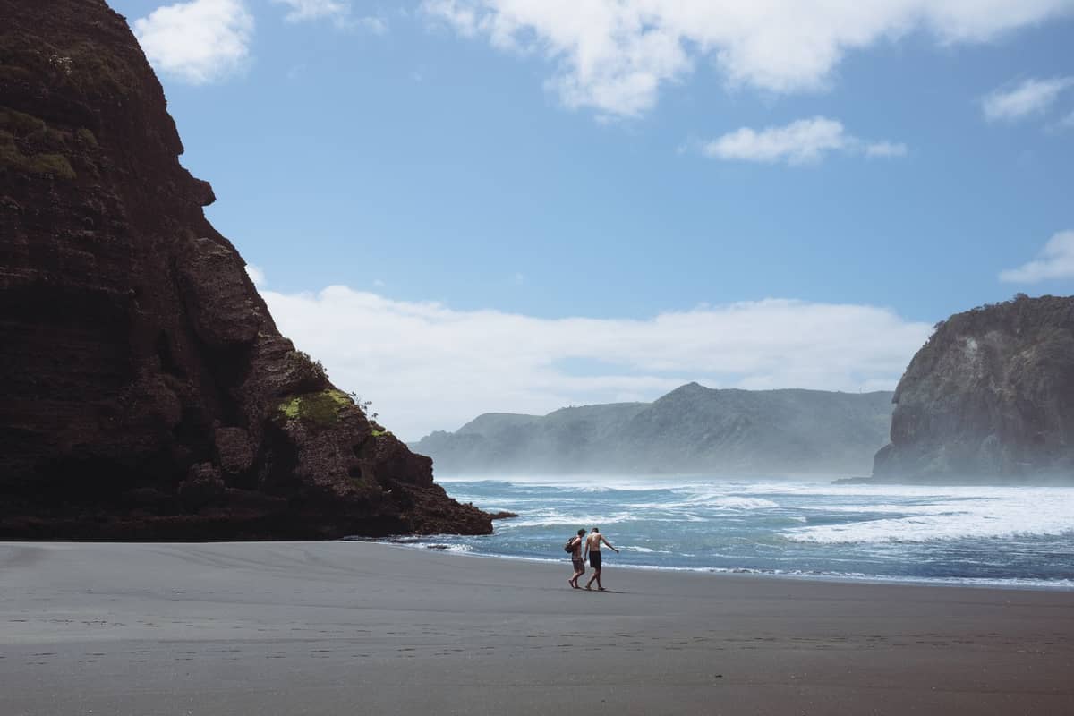 Pareja caminando en una playa de arena negra con acantilados y olas en Karekare, una playa remota de la costa oeste de Auckland