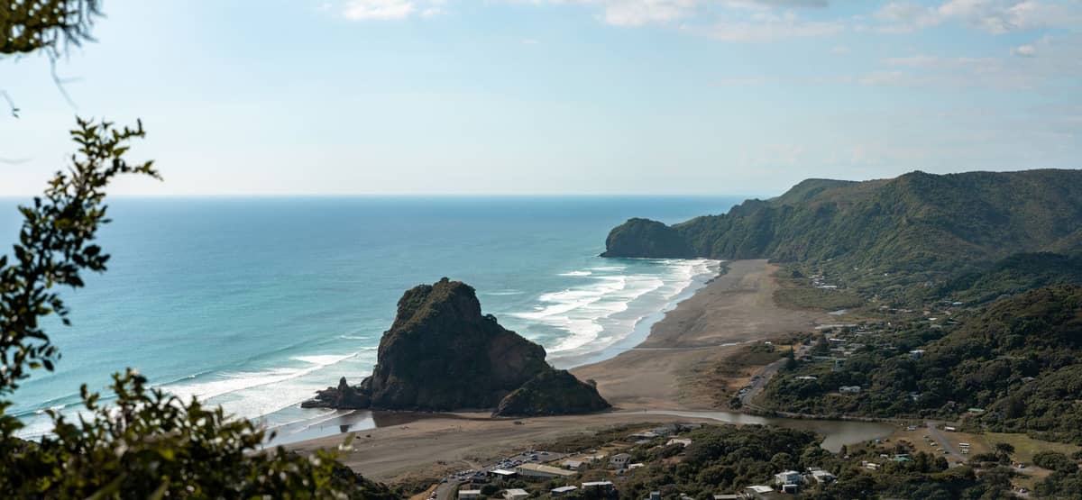 Panoramablick auf Piha Beach und den Lion Rock an Aucklands Westküste mit schwarzem Sand, Wellen und umliegenden Hügeln unter blauem Himmel.