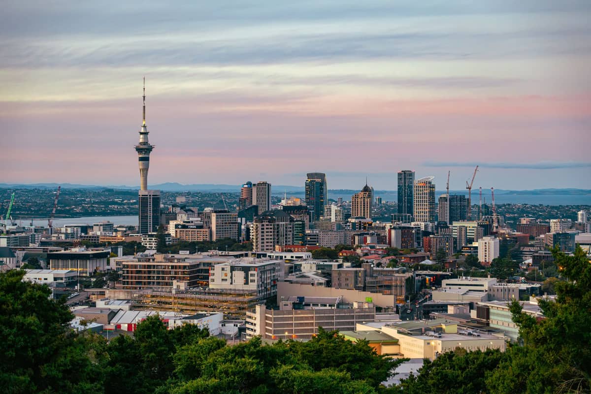 Skyline von Auckland bei Sonnenuntergang mit dem Sky Tower und modernen Stadtgebäuden, eingerahmt von Grünflächen im Vordergrund.