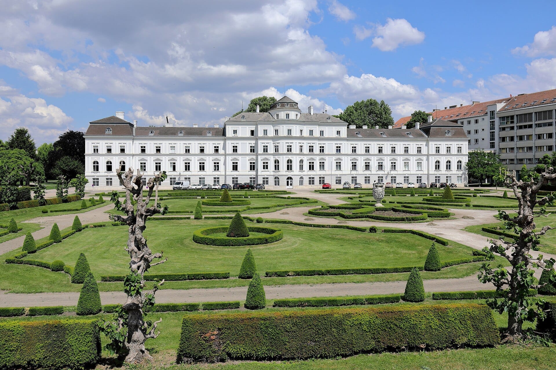 Symmetrische Barockgärten mit gestutzten Hecken und dekorativen Wegen vor dem prachtvollen weißen Palais im Augarten Wien unter blauem Himmel.