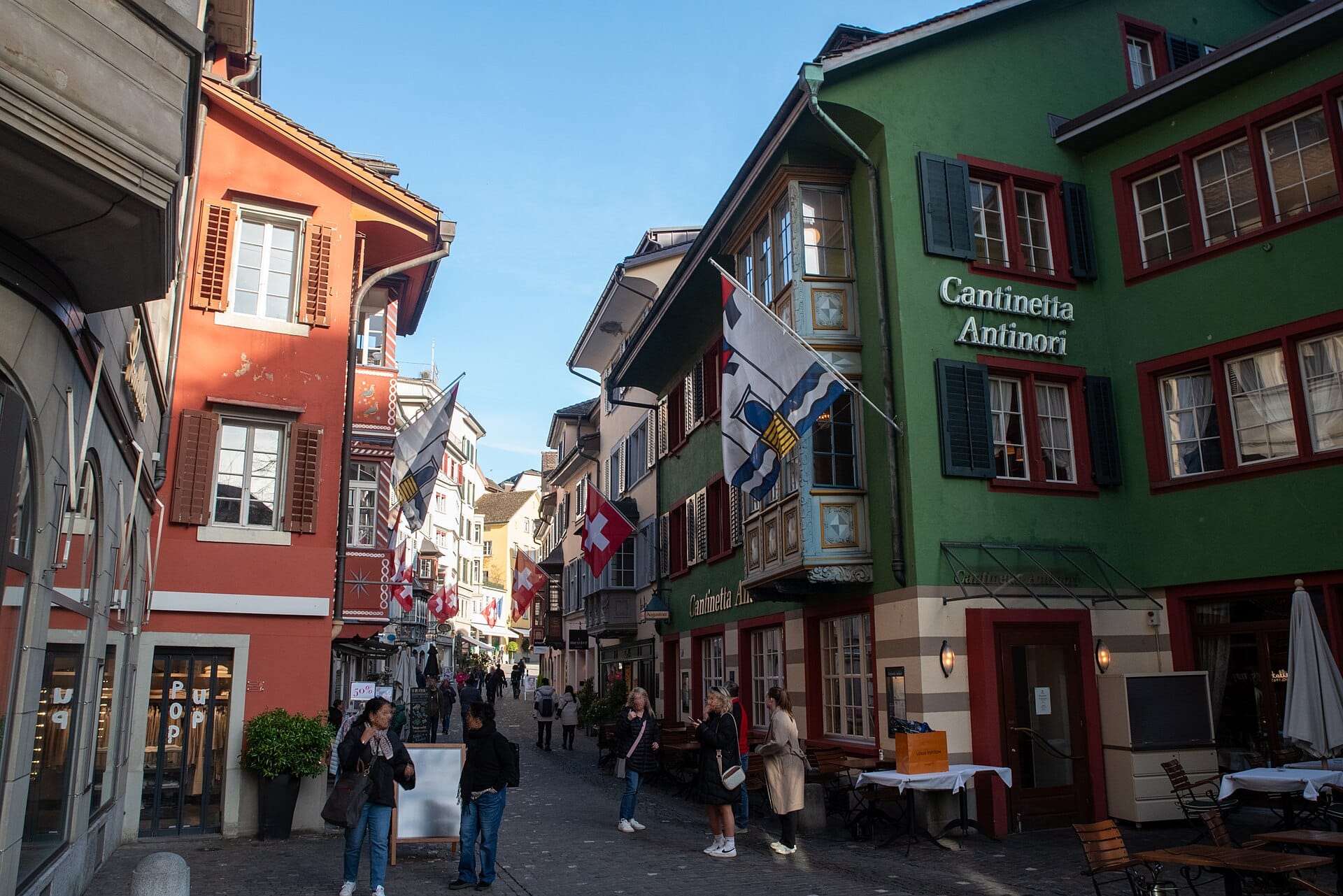 มองยาวไปตามซอย Augustinergasse ในเมืองเก่าซูริก อาคารยุคกลางหลากสี ธงชาติสวิส และผู้คนเดินบนถนนหินกรวดบรรยากาศคลาสสิก