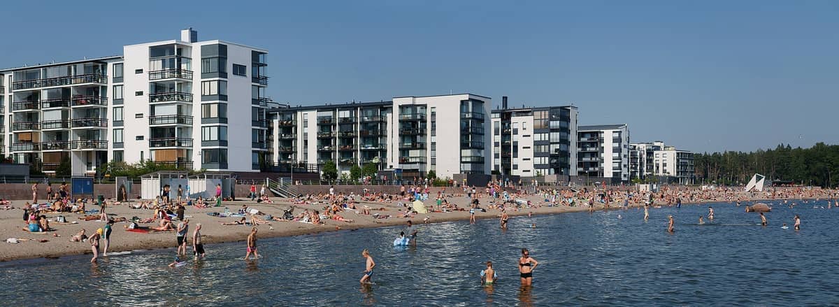 Praia de Aurinkolahti em Helsinki em um dia ensolarado, com modernos edifícios de apartamentos, pessoas tomando banho de sol na areia e nadadores na água.