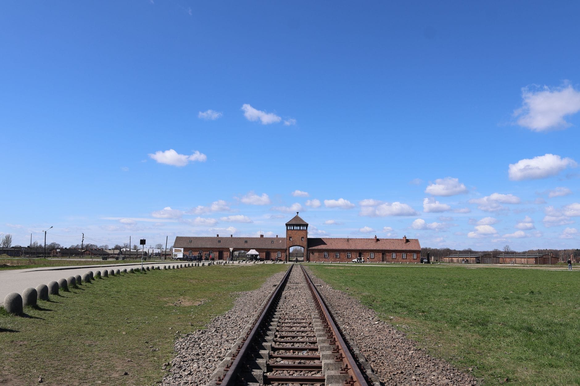 ภาพมุมกว้างของทางเข้าสถานีรถไฟ Auschwitz II-Birkenau ใต้ท้องฟ้าสีฟ้า เห็นประตูแดงอิฐอันโดดเด่นจากระยะไกล