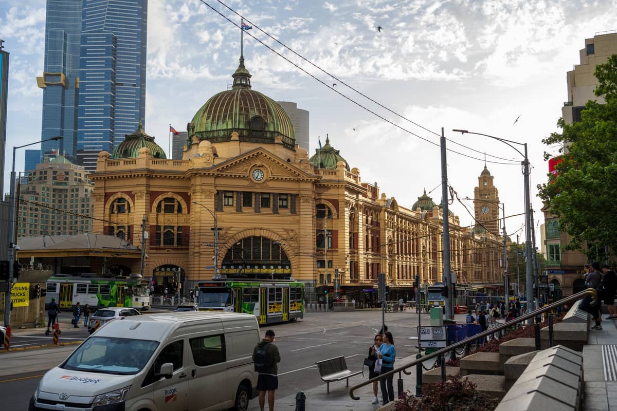 Flinders Street Station im CBD von Melbourne an einem sonnigen Tag, mit Straßenbahnen, Menschen und Wolkenkratzern im Hintergrund.