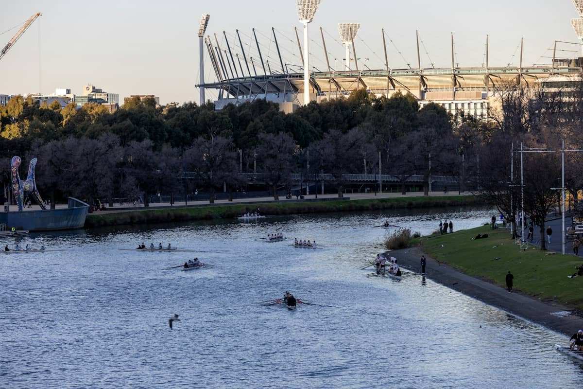 Blick auf das Melbourne Park-Gelände mit dem hinter Bäumen sichtbaren Stadion, dem Fluss im Vordergrund und Ruderern auf dem Wasser.
