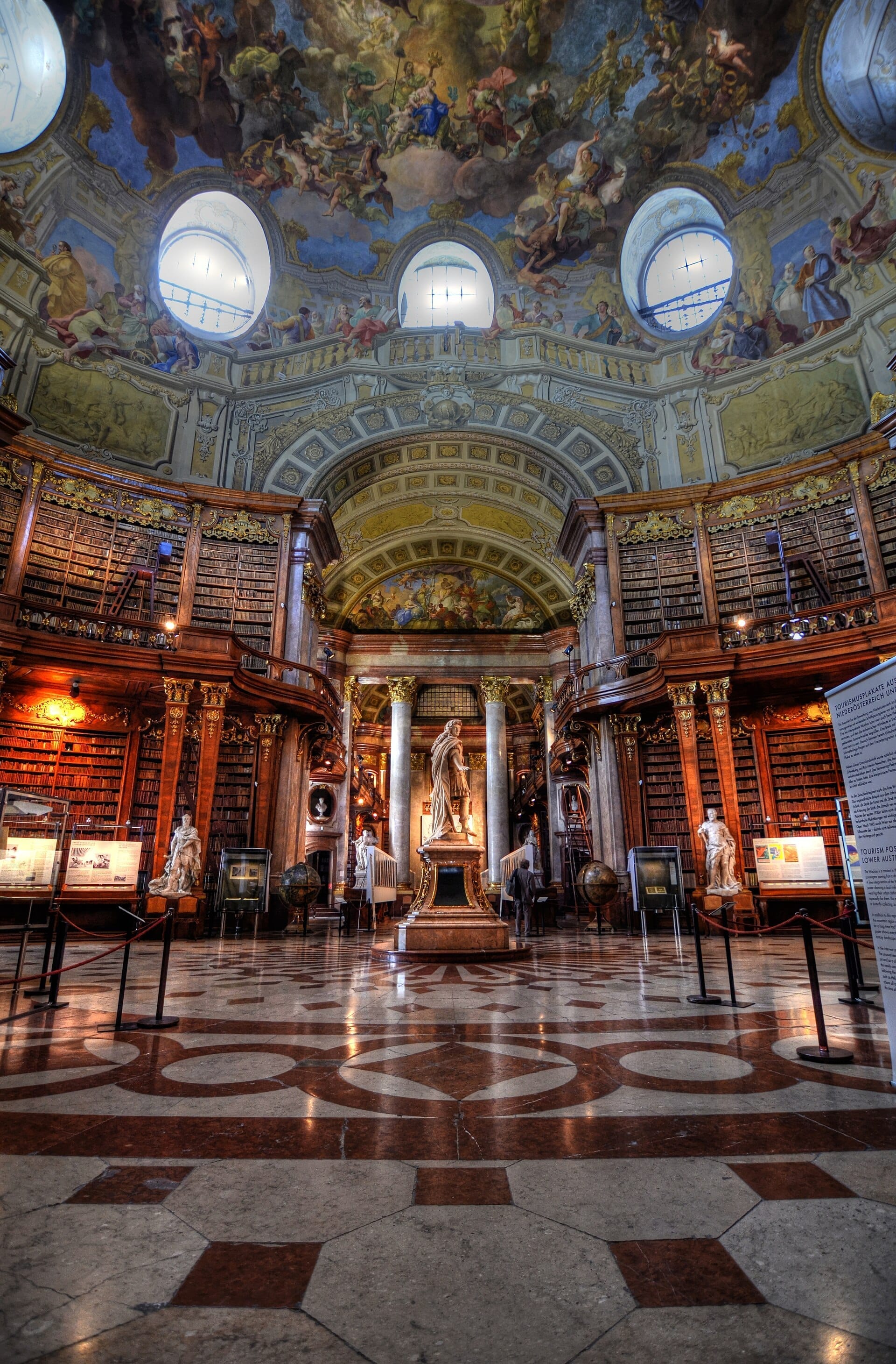 Der Prunksaal der Österreichischen Nationalbibliothek mit Nussbaumregalen, Marmorboden, Statuen und einem prachtvollen barocken Deckenfresko, das von natürlichem Licht erhellt wird.