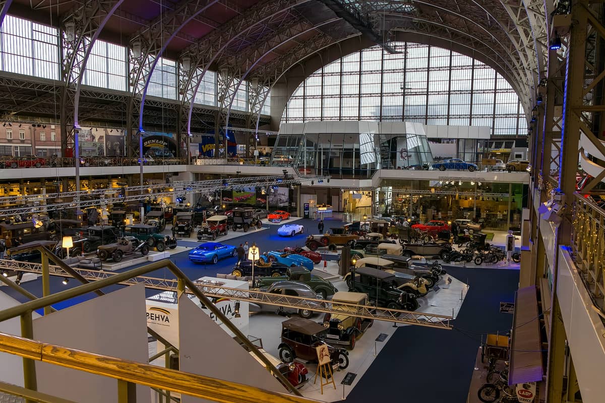 Vista ampla do salão principal de exposições do Autoworld Bruxelas, repleto de carros clássicos sob os altos arcos de ferro e vidro do histórico edifício.