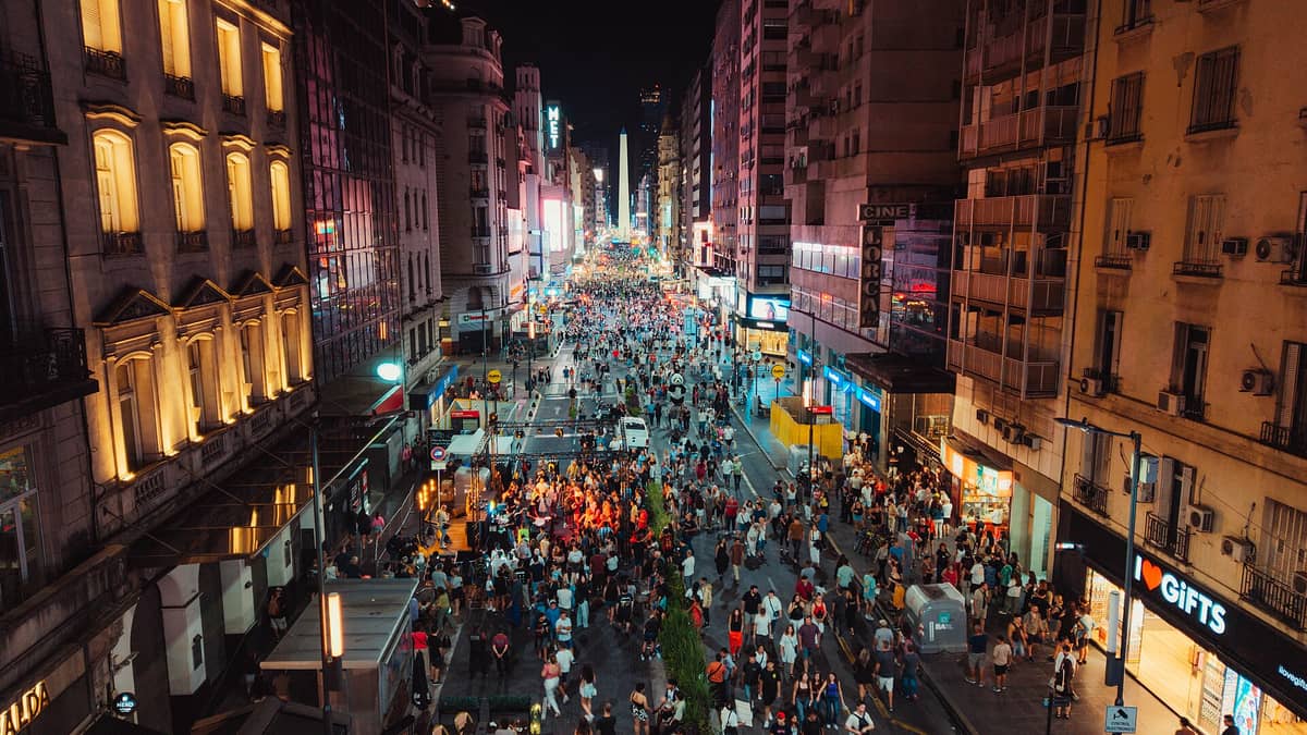 วิวกลางคืนจากมุมสูงของ Avenida Corrientes ในบัวโนสไอเรส เต็มไปด้วยผู้คนและแสงไฟสว่างสดใส ปลายถนนเห็น Obelisco เด่นเป็นสัญลักษณ์