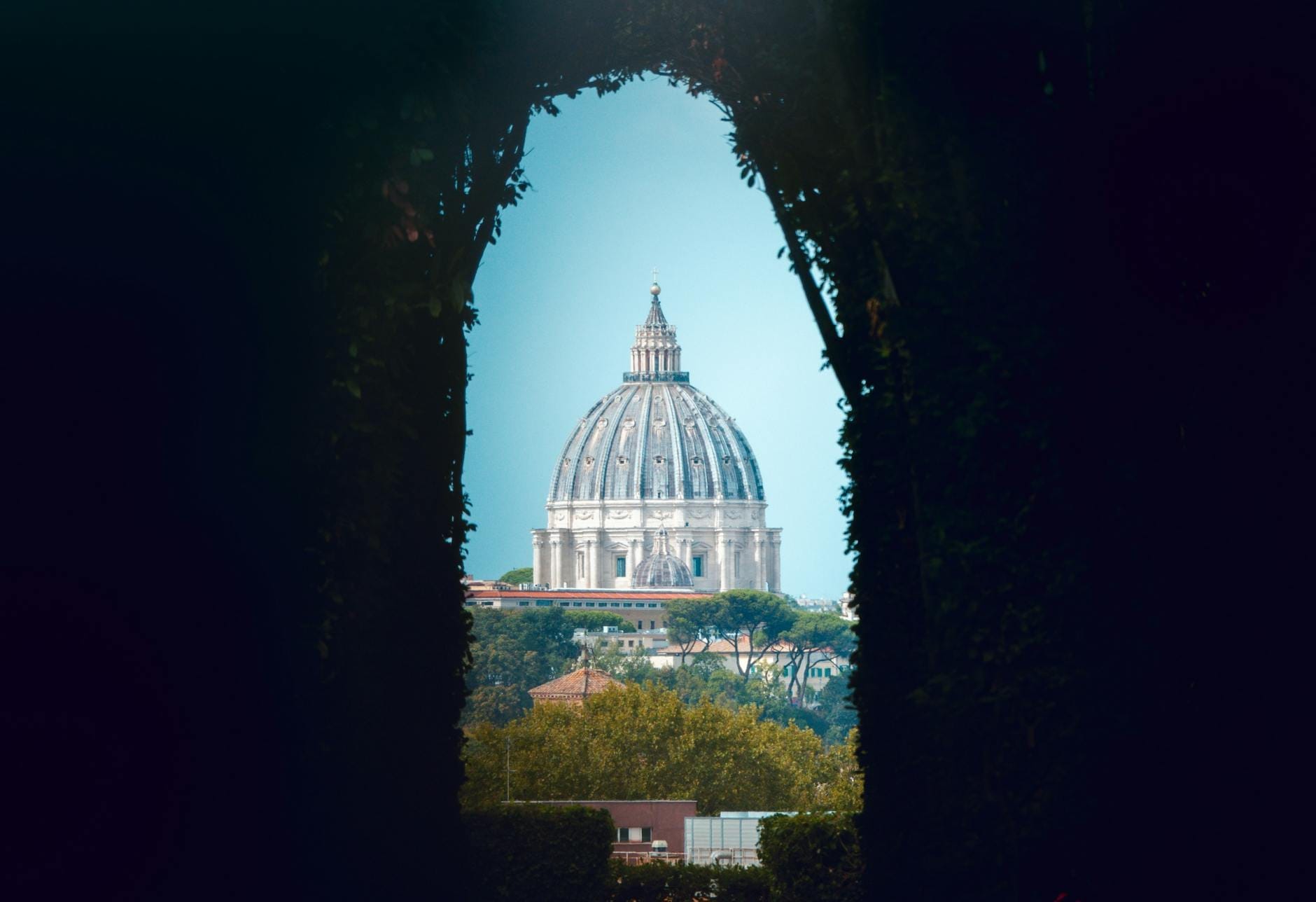 View through the Aventine Keyhole framing the dome of St. Peter's Basilica perfectly, surrounded by dark hedges and bright Roman skyline.