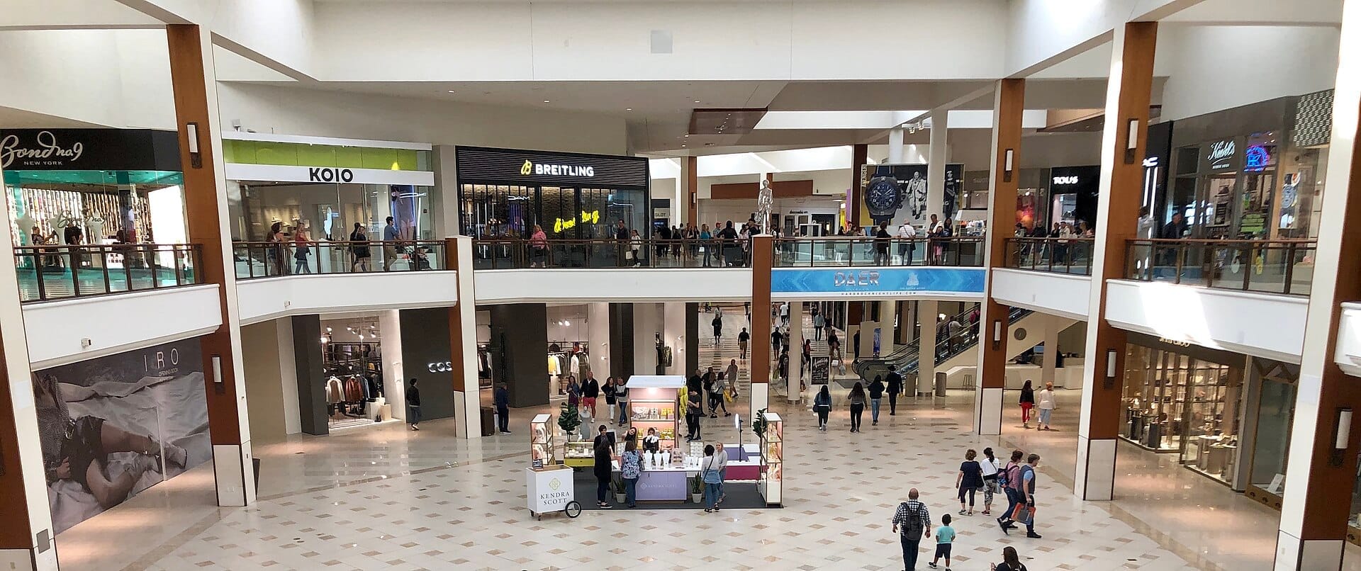 Vista interna spaziosa dell'Aventura Mall con numerosi negozi di lusso, clienti che camminano e un'atmosfera moderna e luminosa con luce naturale.
