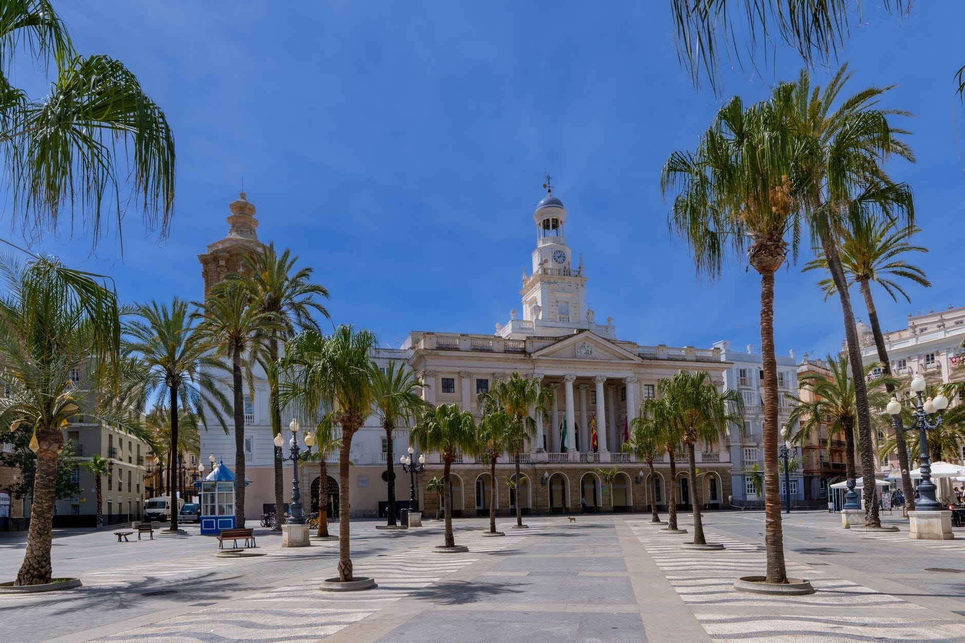 Vista grandangolare dell'Ayuntamiento de Cádiz su Plaza de San Juan de Dios, incorniciato da palme sotto un cielo azzurro brillante, scena di piazza accogliente.