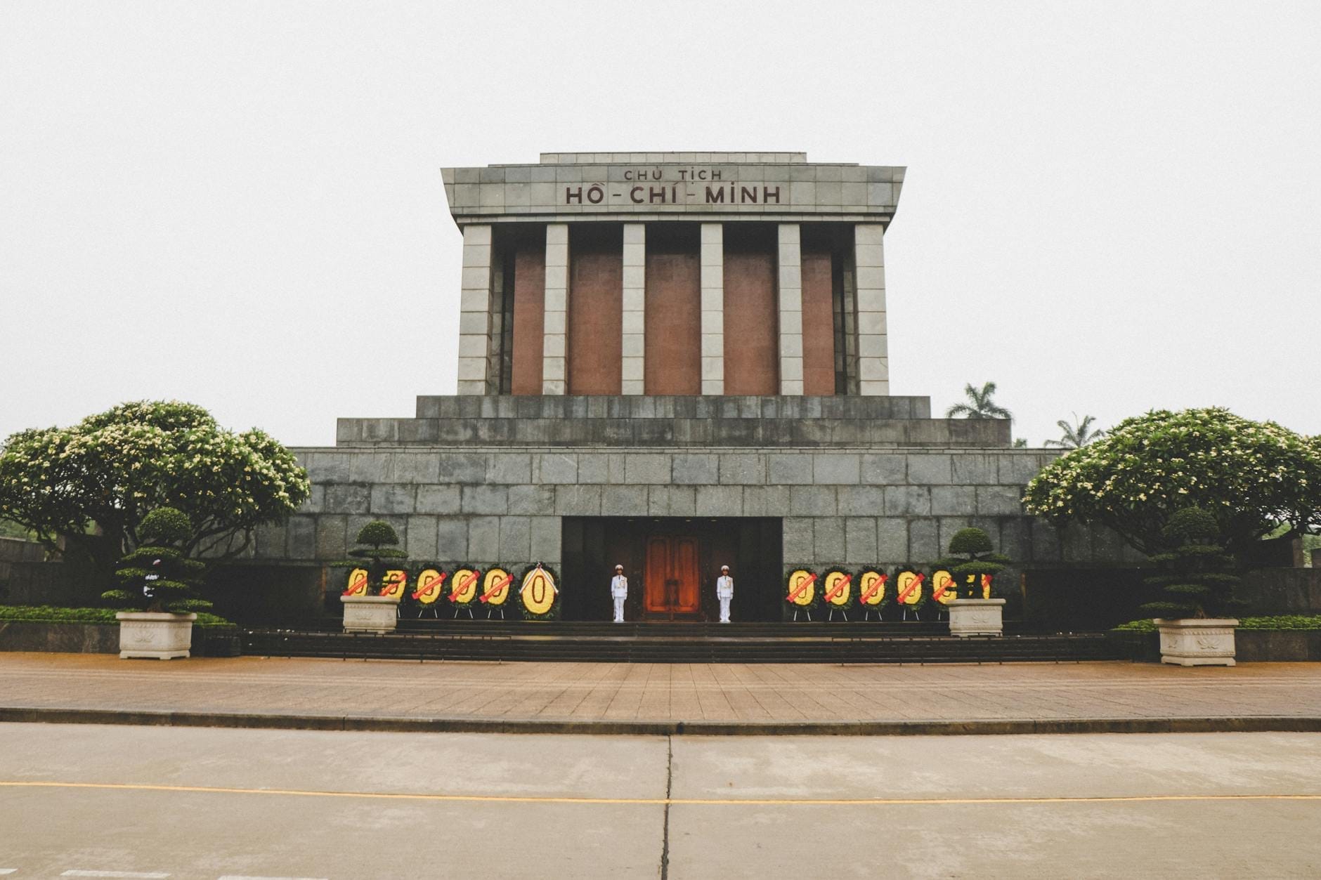 Front view of Ho Chi Minh Mausoleum in Ba Đình with ceremonial guards and flower arrangements, framed by manicured trees and cloudy sky.