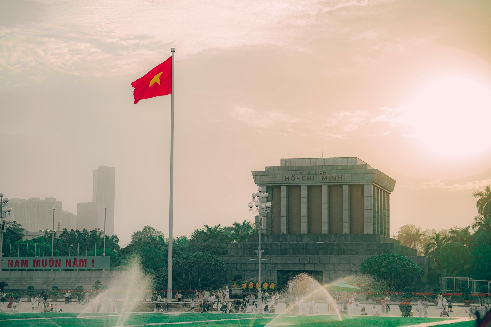Ba Dinh Square at sunset with the Vietnamese flag flying high, fountains in the foreground, and the Ho Chi Minh Mausoleum prominently visible.