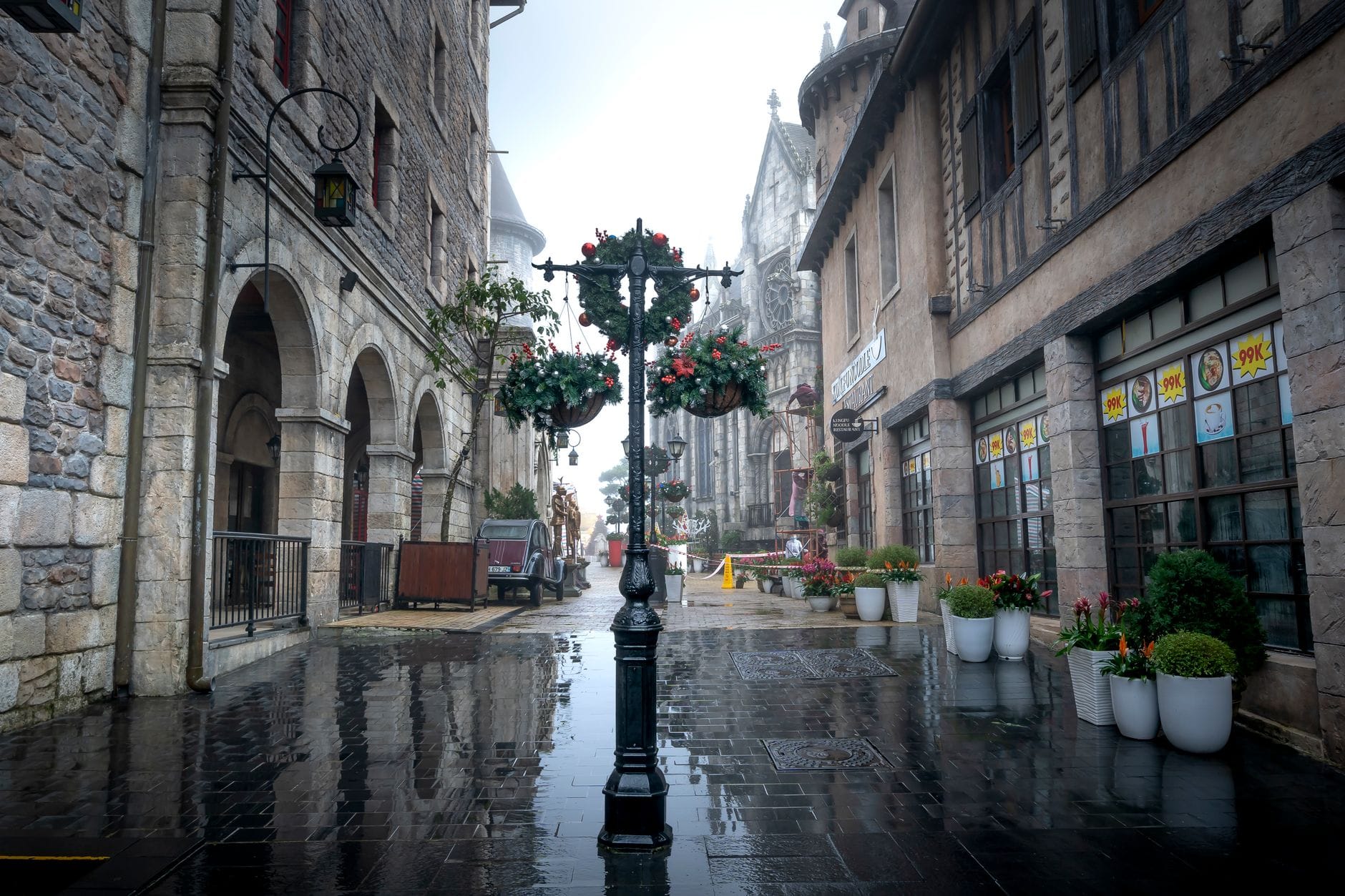 ถนนหินกรวดในหมู่บ้านฝรั่งเศส Ba Na Hills ขณะฝนตก พร้อมป้ายร้านอาหารและอาคารหินที่มองเห็นได้ชัดเจน สะท้อนบรรยากาศพื้นที่รับประทานอาหารในสถานที่