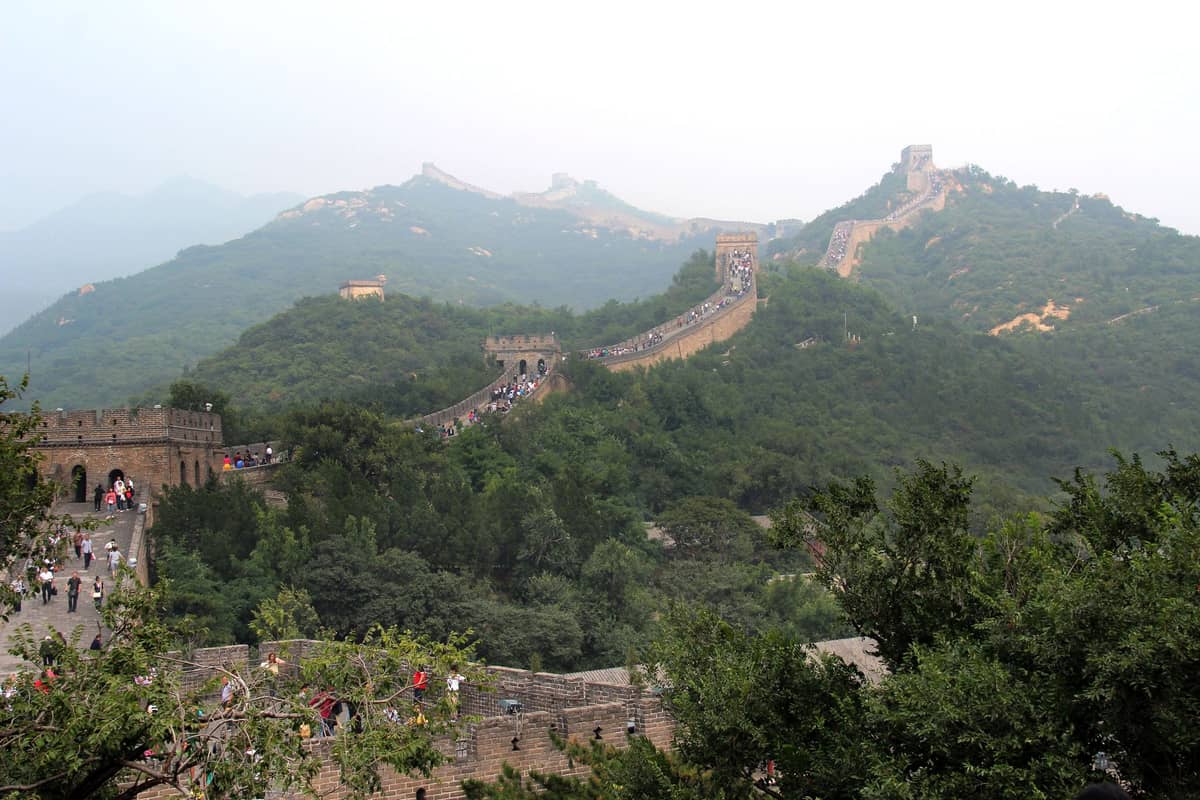 Weitläufiger Blick auf die Chinesische Mauer bei Badaling mit restaurierten Steinzinnen und Türmen, die sich über grüne Hügel schlängeln, an einem diesigen Tag voller Besucher.