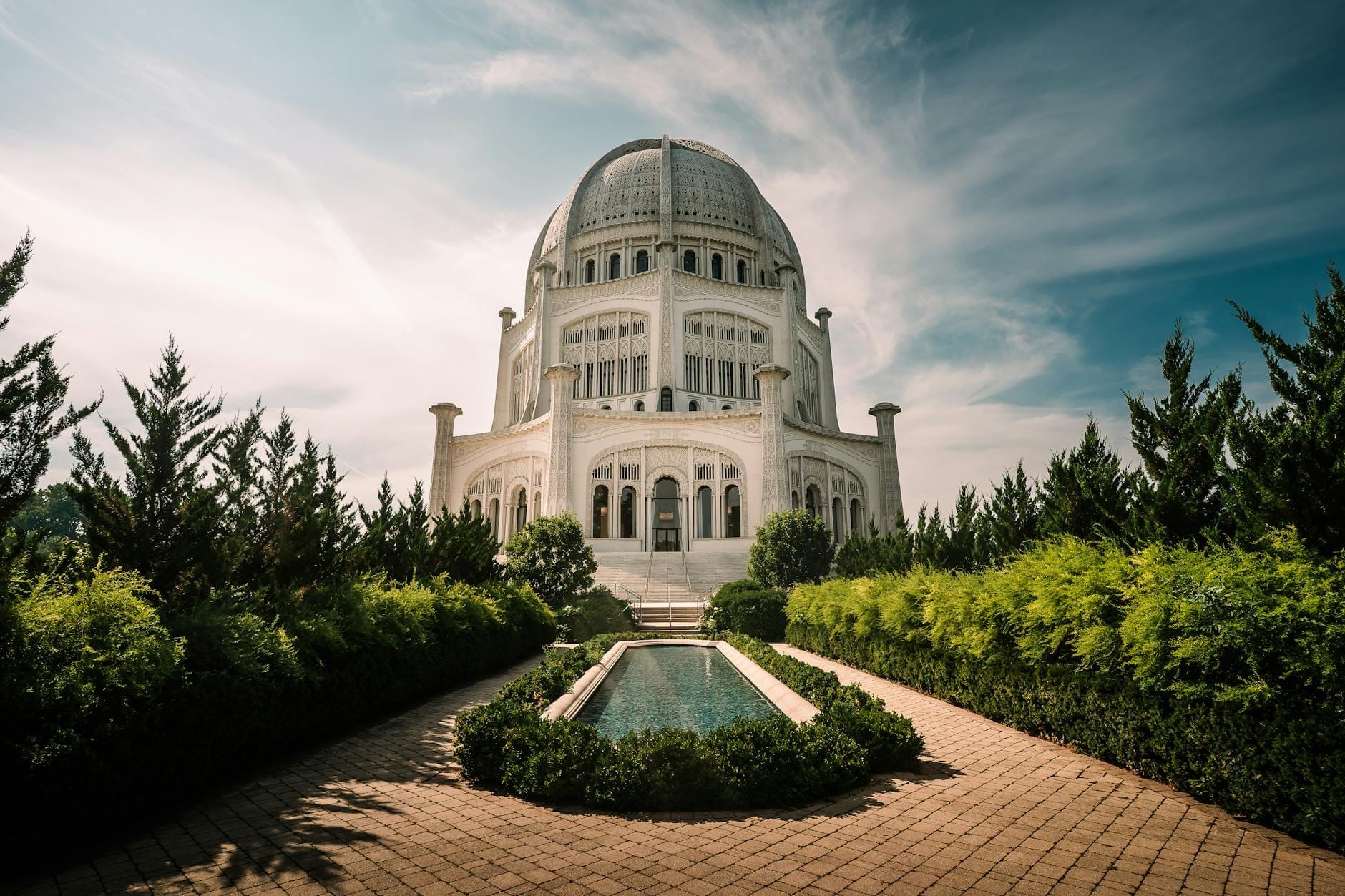 Vista ampia del Tempio Bahá'í di Wilmette, incorniciato da una rigogliosa vegetazione e da una vasca riflettente, sotto un cielo azzurro brillante con nuvole sottili.