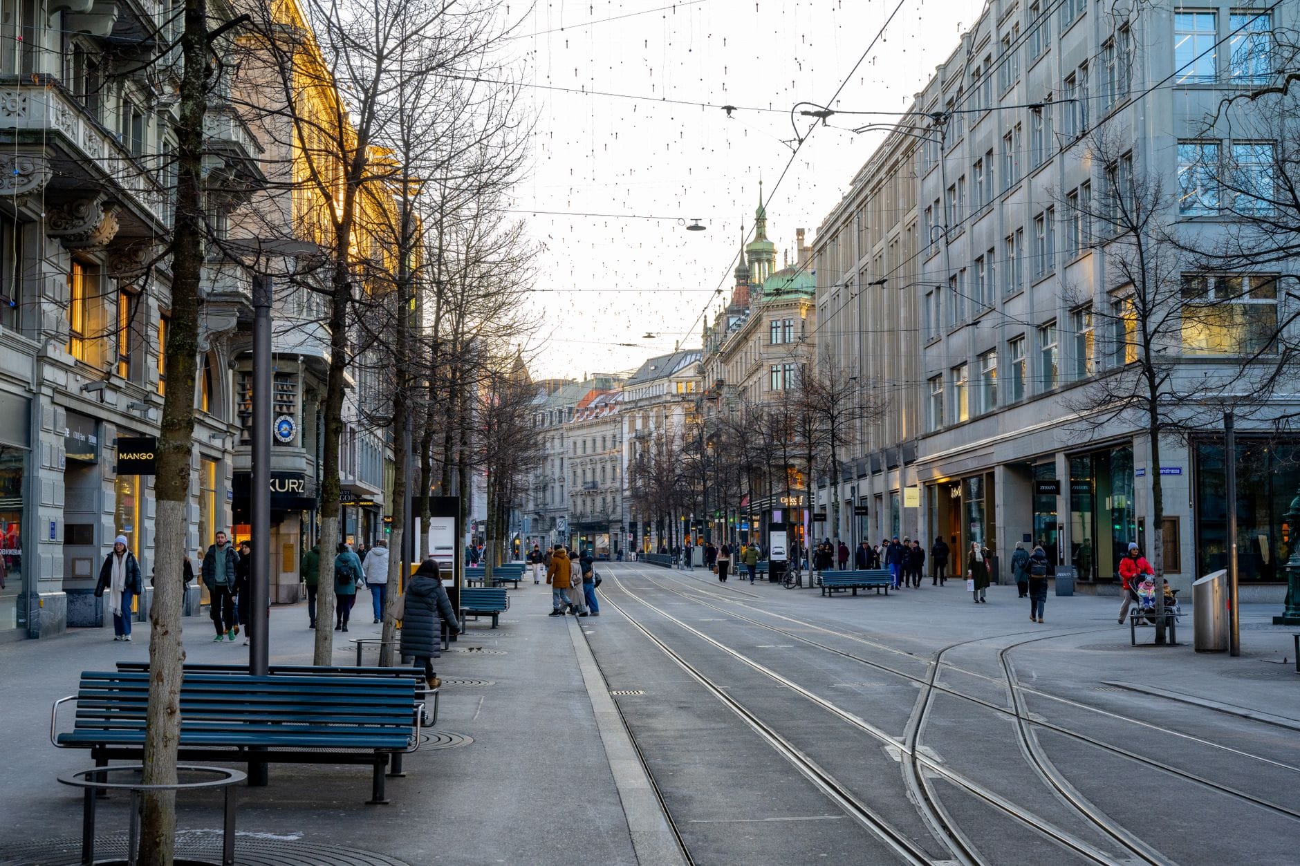 Bahnhofstrasse ซูริคเวลาแดดออก มีรางรถราง ม้านั่ง ร้านค้า และผู้คนเดินพลุกพล่านบนถนนกว้างใจกลางเมือง
