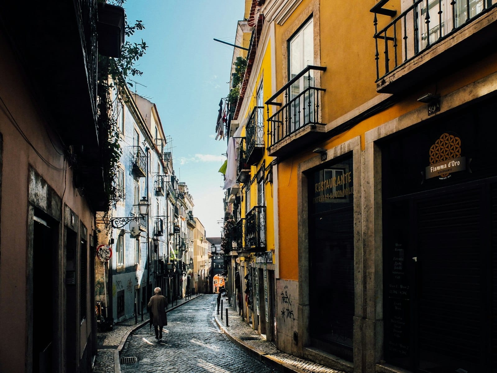 ถนนหินกรวดแคบๆ ใน Bairro Alto ขนาบด้วยอาคารเก่าสีสันสดใสและระเบียง มีผู้คนเดินอยู่ไม่กี่คนใต้แสงกลางวันที่สว่างจ้า