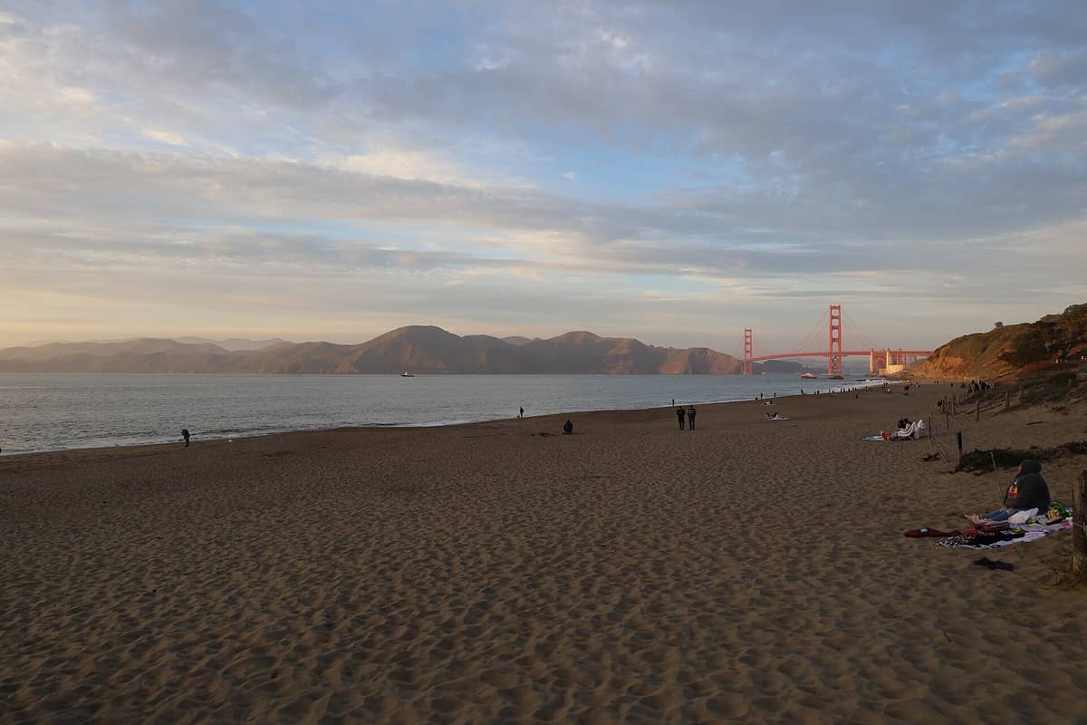 Weiter Sandstrand des Baker Beach bei Sonnenuntergang mit einzelnen Menschen in der Ferne, Wellen und der Golden Gate Bridge unter einem teils bewölkten Himmel.