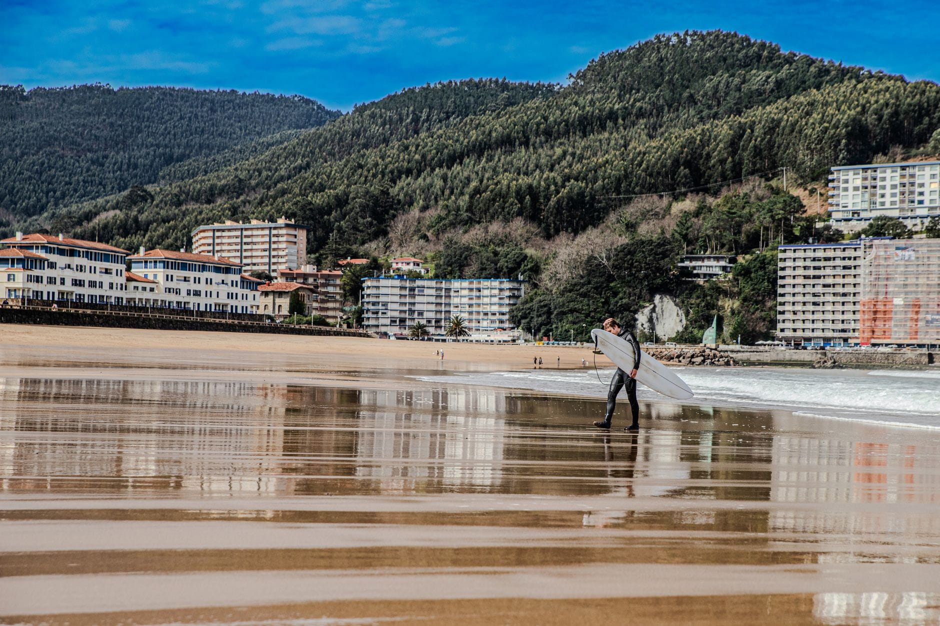 Ein Surfer läuft über den nassen Sand von Bakio Beach, während Wellen, der Ort und saftig grüne Hügel unter klarem Himmel im Hintergrund zu sehen sind.